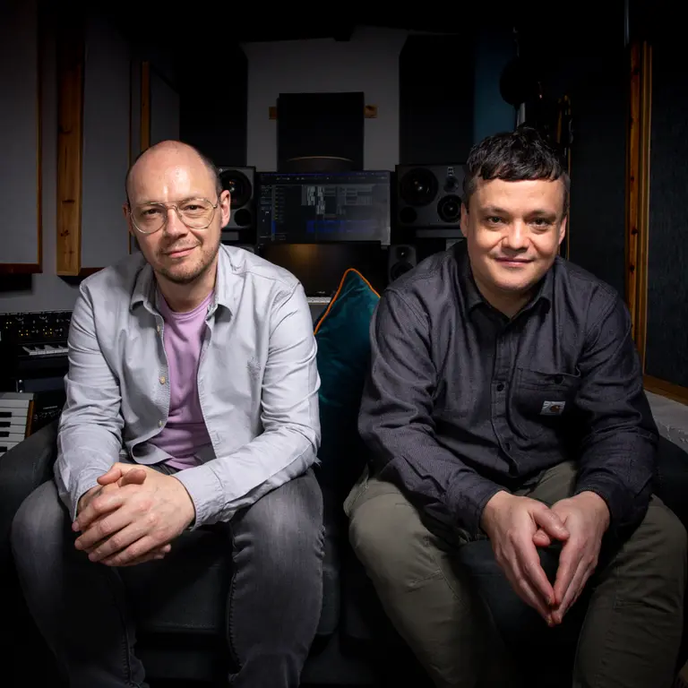 Two men sit on a sofa and smile at the camera