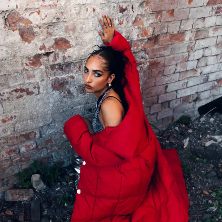 Roukaya B wearing a red jacket against a brick wall, turning and facing the camera