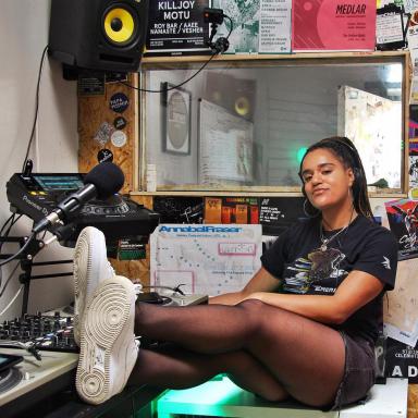 Young person with long braids, wearing all black with white trainers and sitting in a studio with her feet resting on the table.