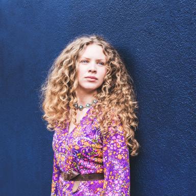 Young woman with long curly hair and wearing a purple patterned dress.