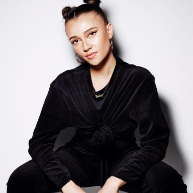 Woman wearing all black, with hair in two buns, sitting down with legs spread, against a white background.