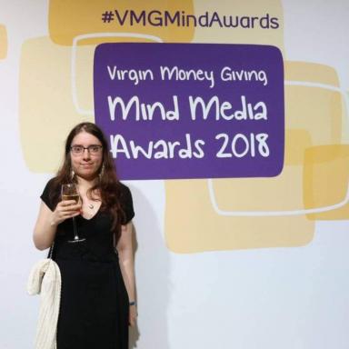 Young person with brown hair, wearing a black dress, standing in front of a poster that says "Mind Media Awards 2018".