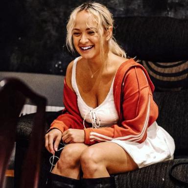 Candid photo of young person laughing, with blonde hair and wearing a white dress, orange jacket and black boots.