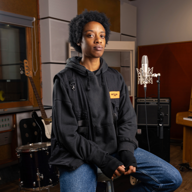 a person with short curly hair sits on a stool in a music studio